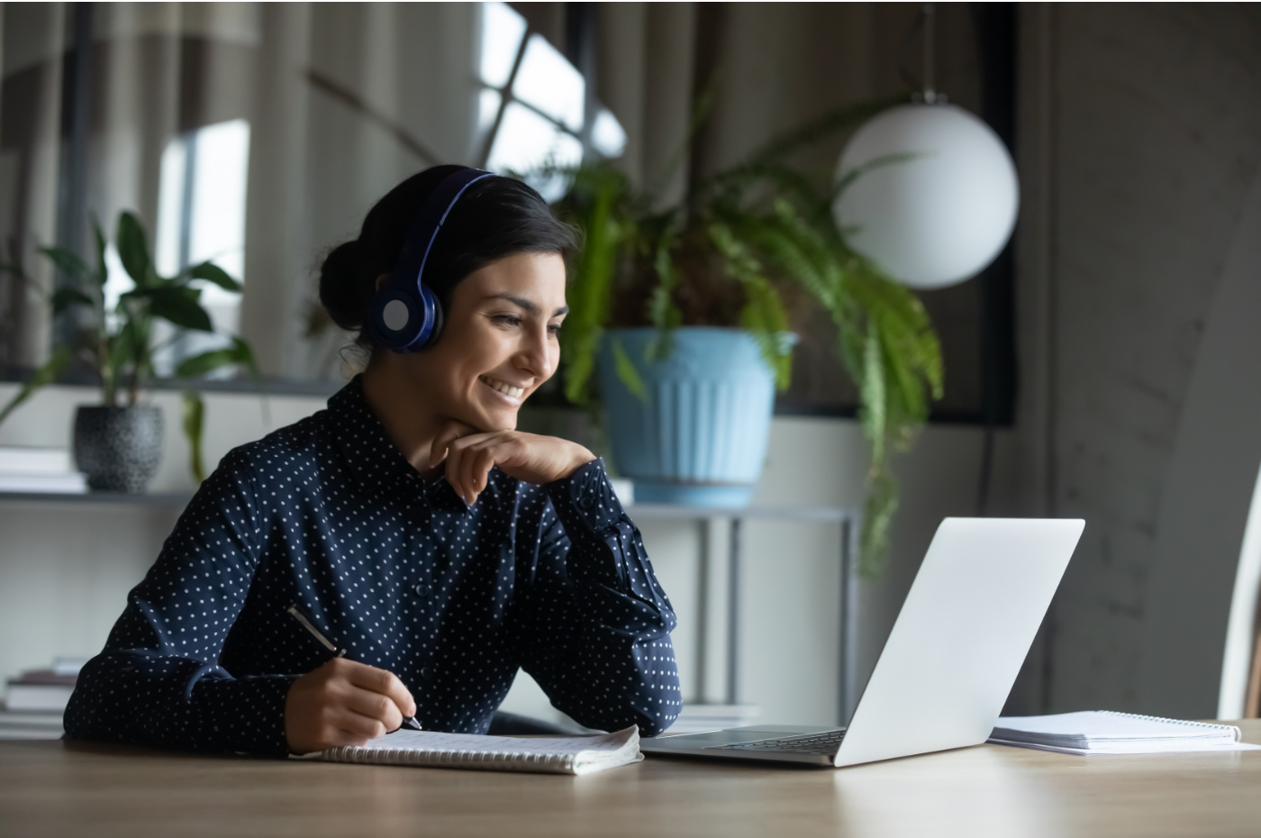 Woman sitting at table, working on computer and wearing a headset.